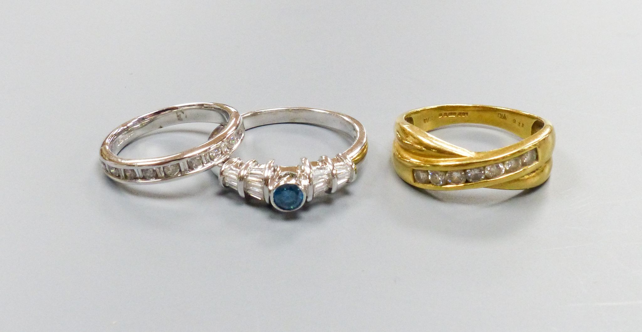 Three modern dress rings including a 750 yellow metal and seven stone channel set diamond crossover ring, size O, an 18ct white gold and nine stone diamond set half eternity ring, size I and a 750 white metal and blue di